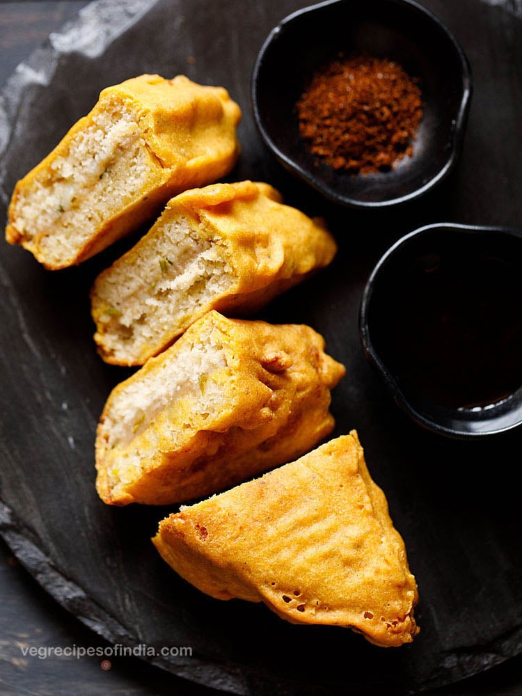 stuffed bread pakoda slices on a black slate board with two black small bowl of dry garlic chutney and tamarind chutney
