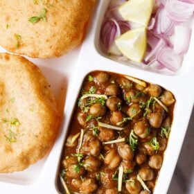 closeup shot of white plate with sections filled with street style chole masala, sliced onions, fresh lime wedges and bhatura on a marble table-top