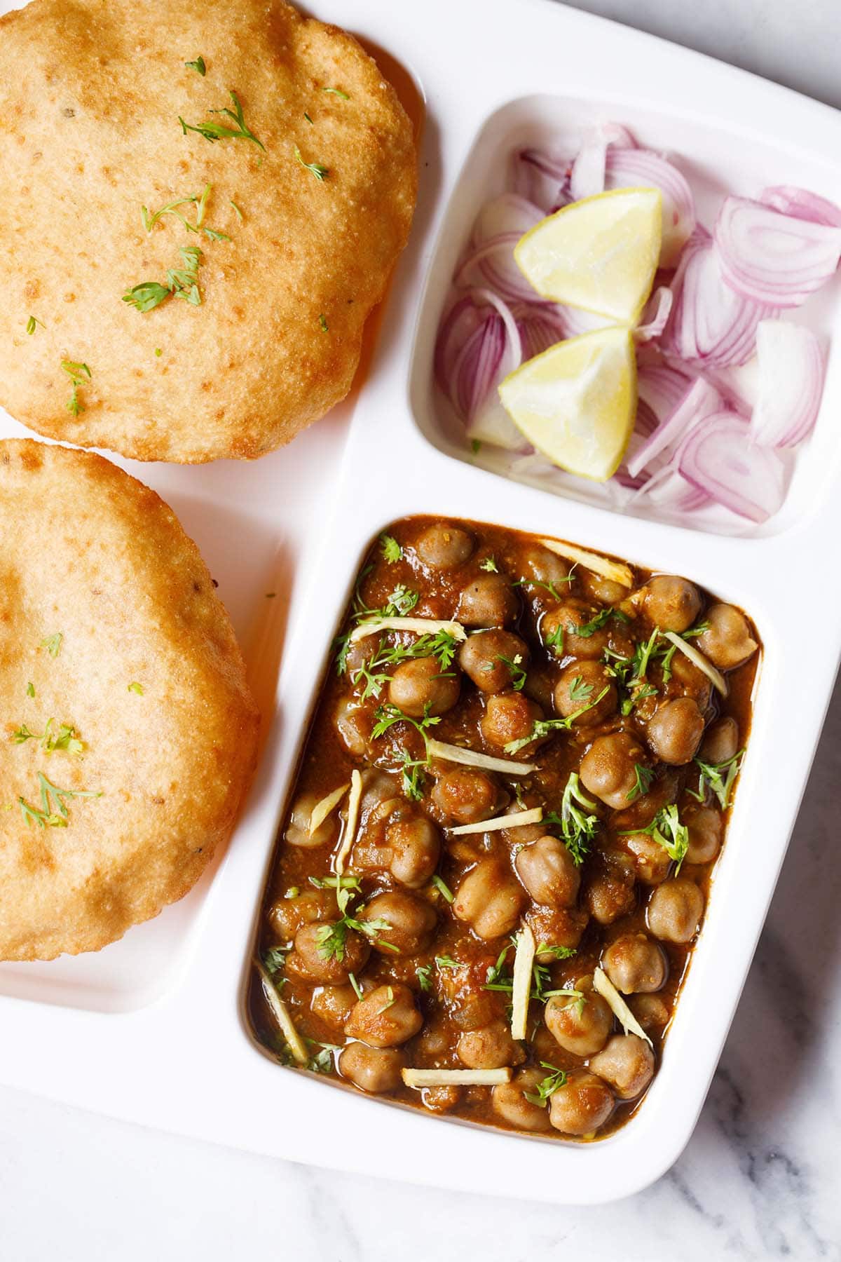 closeup shot of white plate with sections filled with street style chole masala, sliced onions, fresh lime wedges and bhatura on a marble table-top