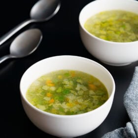 clear veg soup in two white bowls with two spoons on left side on a black table.