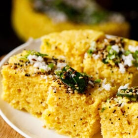 45 degree angle shot of khaman dhokla squares on a white plate garnished with cilantro, curry leaves and the fried curry leaves and spices mixture