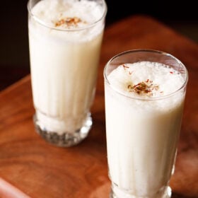 sweet lassi topped with crushed cardamom powder and saffron in two tall glasses on a wooden board