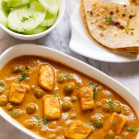 Top shot of matar paneer on white rectangle plate next to sliced cucumber in a white bowl and folded two roti in a white plate sitting on white marble table