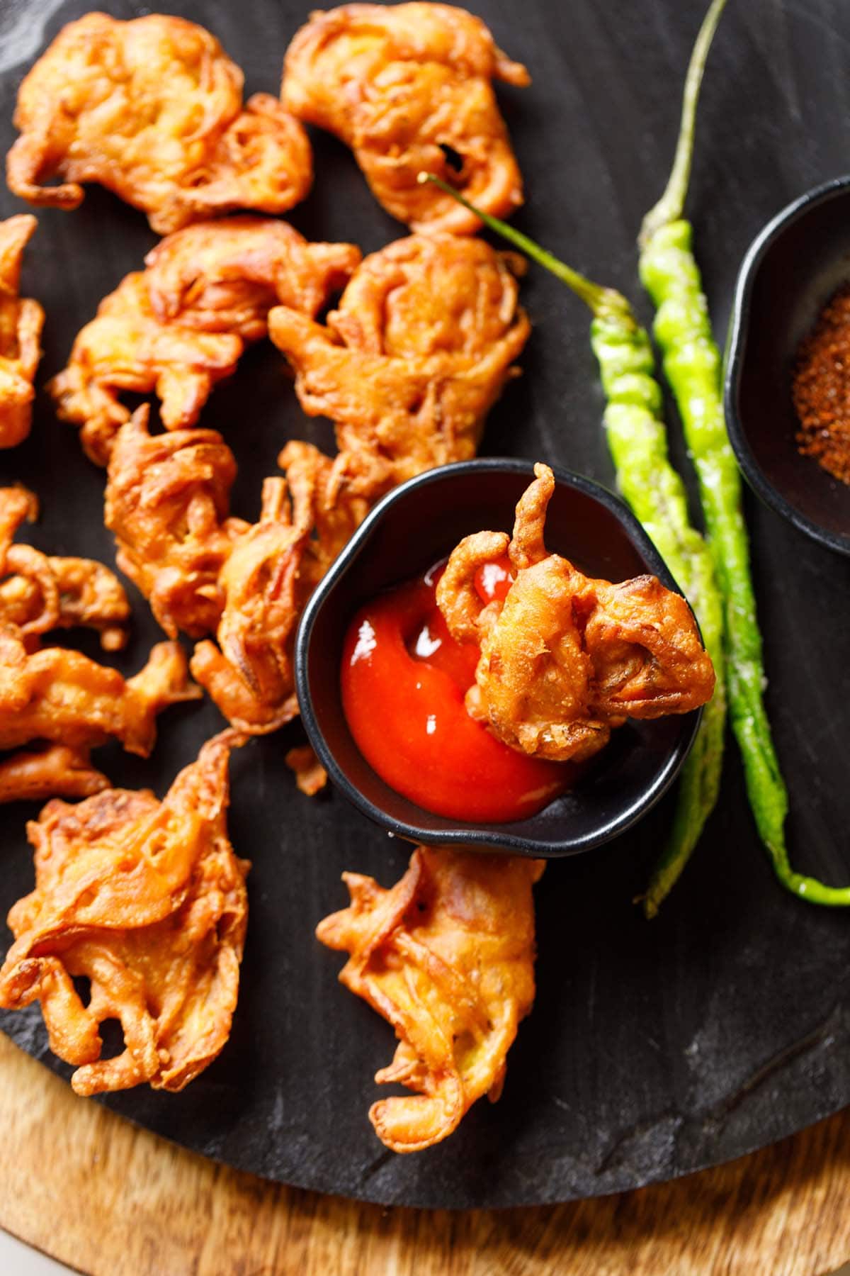 onion pakoda served on a black granite platter with tomato ketchup and fried green chilies. 