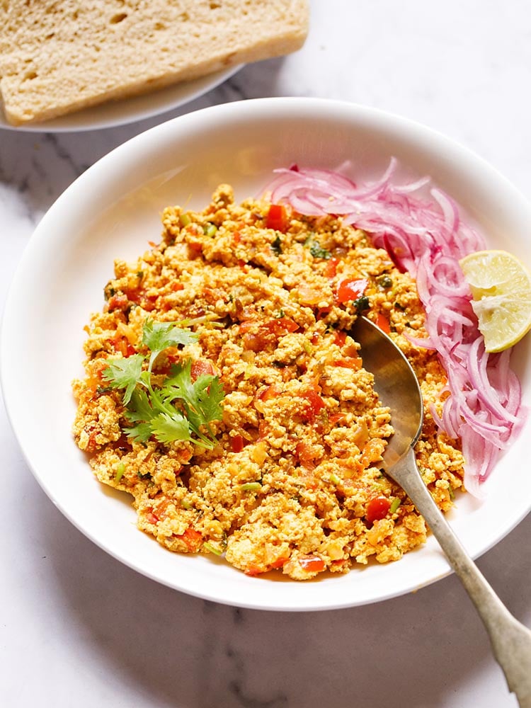 Paneer Bhurji served in a shallow white bowl garnished with one coriander sprig with a spoon inside the bhurji and topped with some sliced onions and a wedge of lemon
